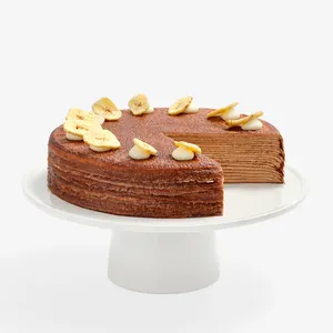 A whole chocolate Mille Crêpes cake on a white pedestal stand with a slice removed, showing thin cocoa-colored crepe layers inside. The top is garnished with dollops of white cream and dried banana slice toppings along the rim.