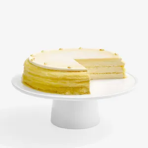 A whole pale-yellow Mille Crêpes cake on a white pedestal stand with a slice removed, revealing thin cream-colored layers inside. The top is covered with a smooth light glaze and garnished around the edge with small gold leaf accents.
