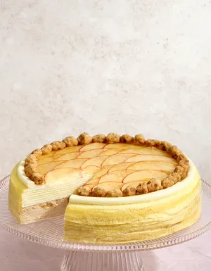 A golden Mille Crêpes cake on a clear ribbed glass stand with a slice removed, revealing two distinct internal cream and filling layers. The top is decorated with thinly sliced fruit arranged in a fan-like pattern, bordered by small golden-brown crumble clusters and a smooth white cream ring, set against a light pink textured background.