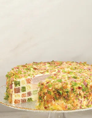 A whole cake covered in vibrant multicolored flakes on a clear glass stand, shown alongside a slice served on a gold-rimmed white plate with a gold fork and napkin. The cut section reveals a unique interior combining thin pastel crepe layers and a colorful checkerboard pattern of cake squares, set against a light marble background