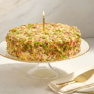 Lady M Birthday Cake 9" covered in colorful crêpefetti with a lit gold candle on a cake stand