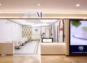 Storefront entrance of a brightly lit Lady M boutique featuring an illuminated 'LADY M' sign overhead. The interior features curved ceiling lighting, white dining chairs and small tables over patterned floor tiles, and a fluted service counter with a refrigerated cake display case. To the right stands a large vertical digital screen displaying a cake slice and store location text.